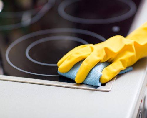 people, housework and housekeeping concept - hand in rubber glove with rag cleaning cooker at home kitchen